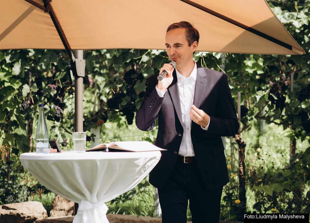 Der freie Redner Oliver MEtzger steht an einem Stehtisch unter einem Sonnenschirm und spricht bei einer freien Trauung